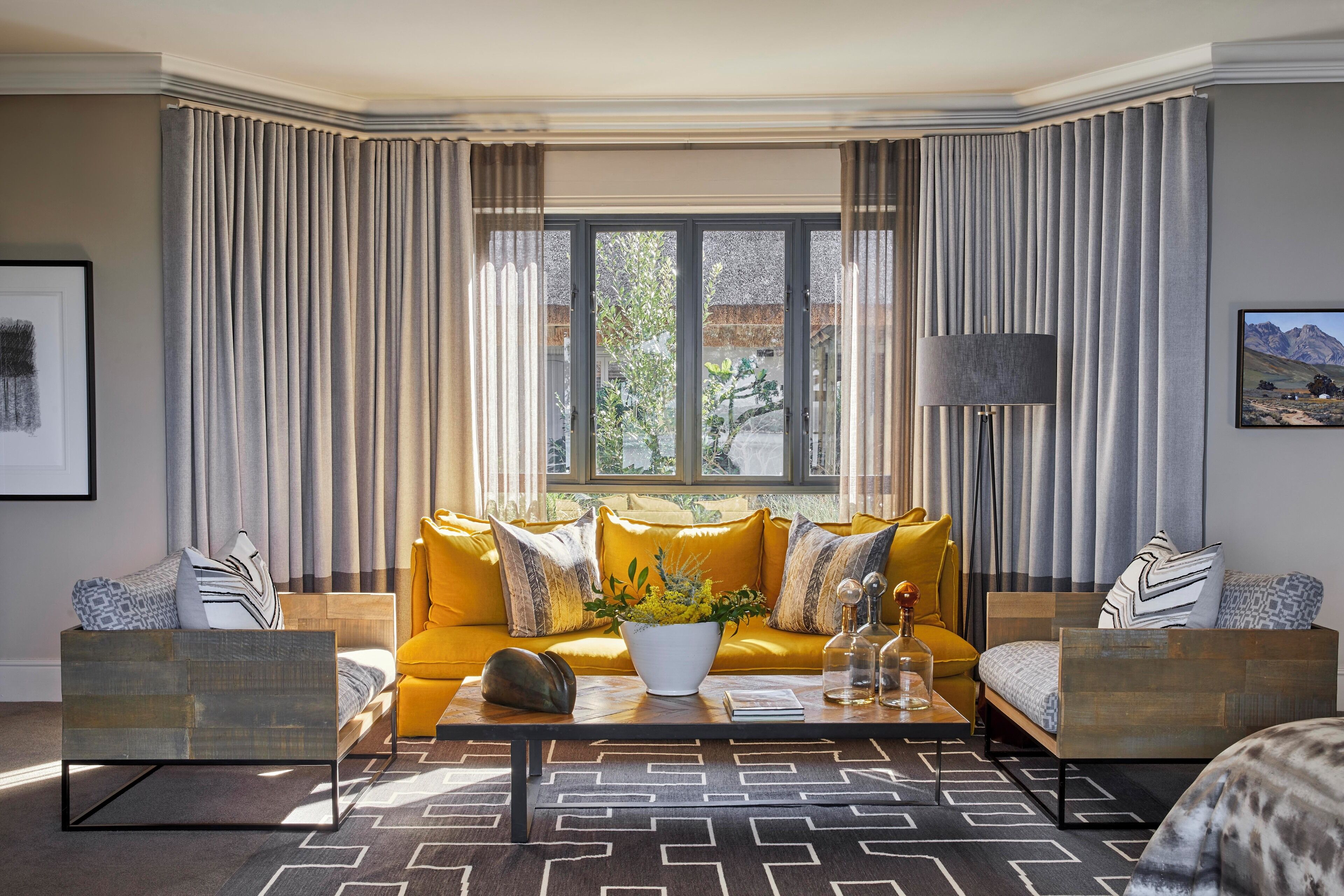 A modern living room with a yellow sofa, a coffee table with a vase, and a view of greenery through large windows.