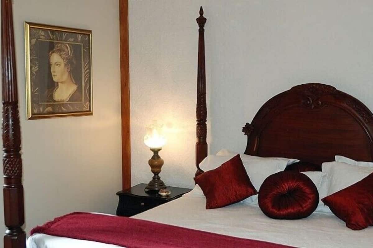 Una cama con dosel, un tablero de cabecera de madera, una mesita de noche con lámpara y un retrato enmarcado en la pared.
