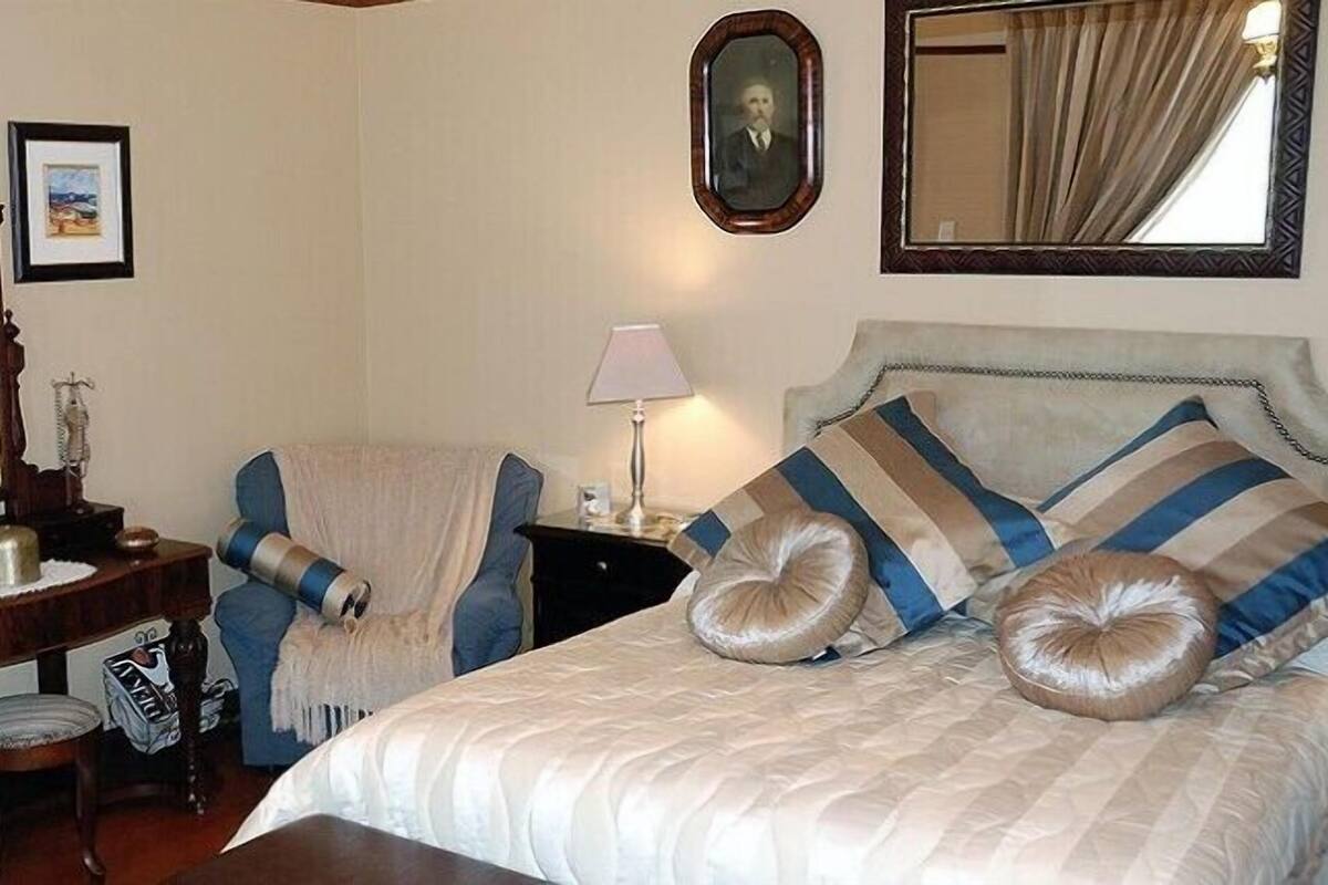 Un cuarto de hotel con una cama, dos sillones, un tocador con espejo, una lámpara y cuadros en la pared.