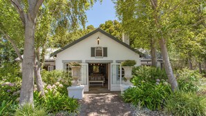 A quaint, single-story house with a brick pathway leading to the entrance, surrounded by lush greenery and trees. - Rose Cottage (Franschhoek)