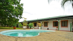 Uma área de piscina com um terraço de tijolos, rodeada por plantas em vasos e um edifício com telhado verde.