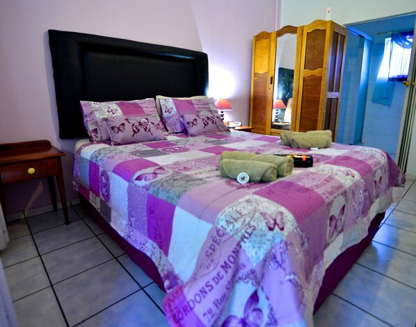 Una cama bien hecha con colcha y almohadones estampados, una mesita de noche con lámpara y un armario de madera al fondo.