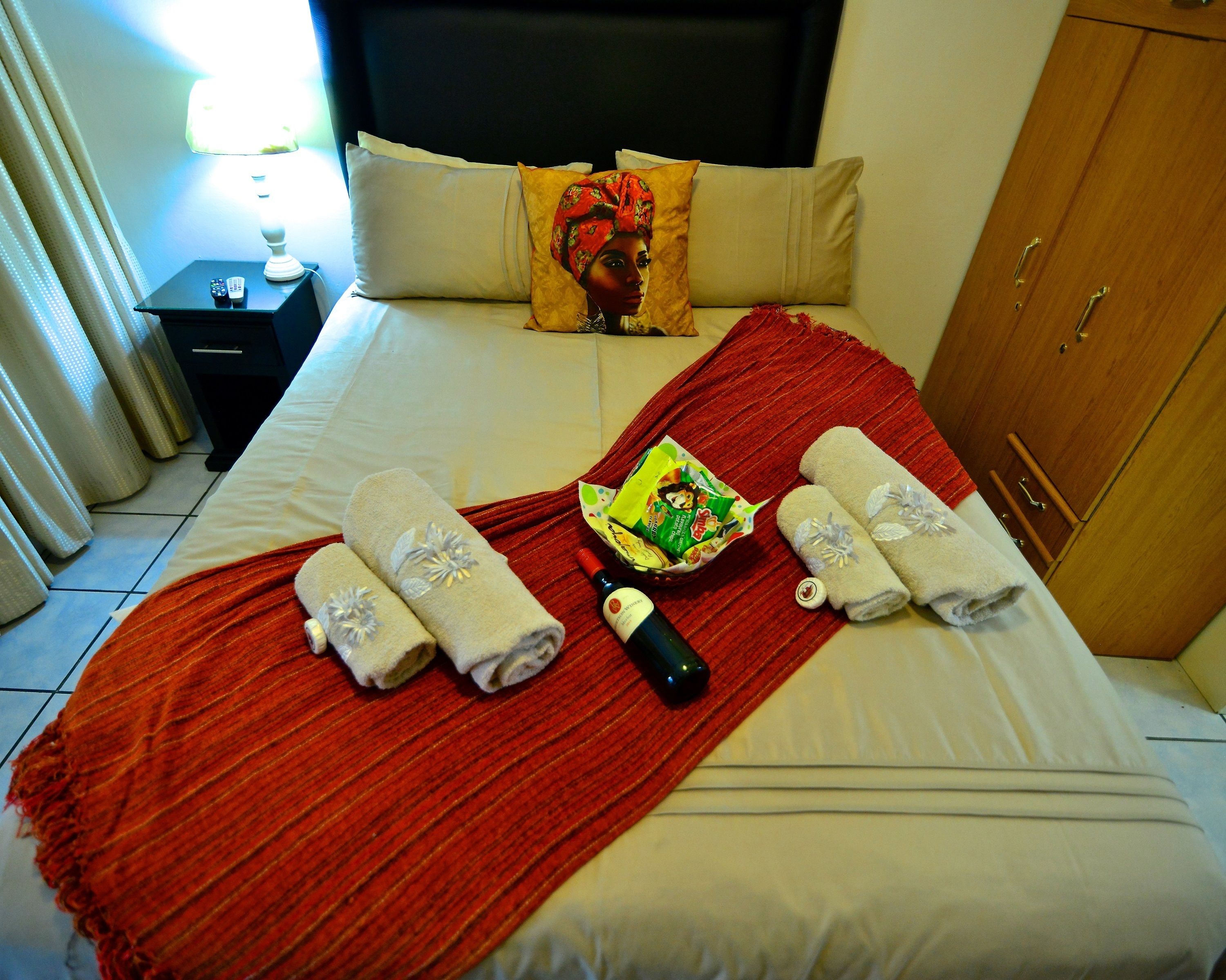 A hotel room with a bed, a red throw blanket, towels, a bottle of wine, and a small table with a remote control.