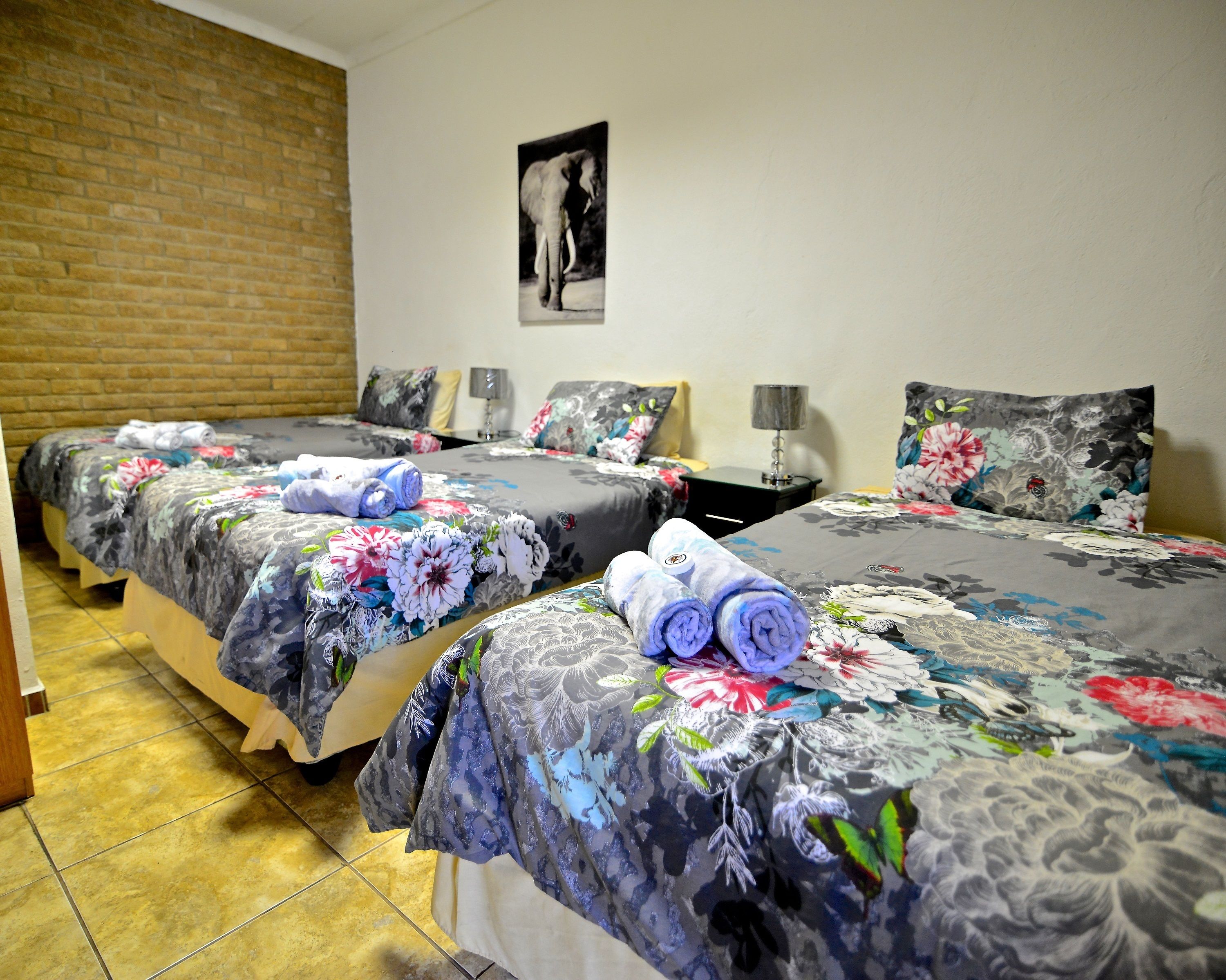 A hotel room with two beds, each with floral bedding and rolled towels. There is a brick wall and a framed picture on the wall.