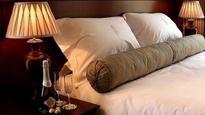 Una habitación de hotel con una cama bien hecha, dos lámparas de noche, una botella de vino y dos copas sobre una mesita.