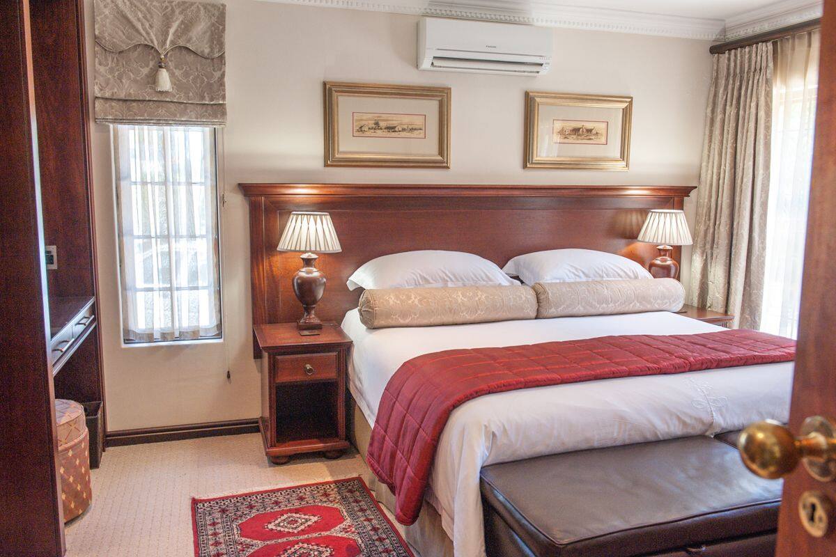 A hotel room with a large bed, two bedside lamps, a nightstand, a wooden headboard, a window with curtains, and two framed pictures on the wall.