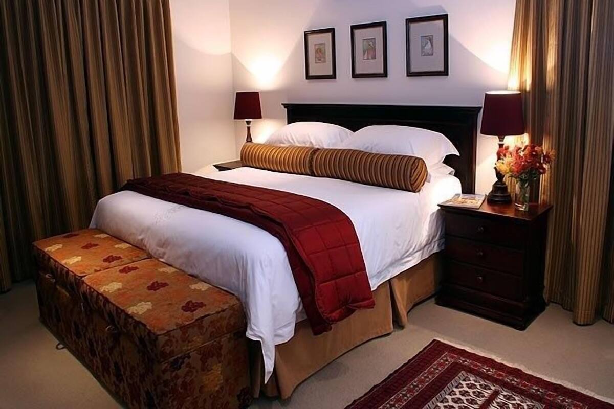 A hotel room with a bed, a nightstand, a lamp, a vase with flowers, and a red and white patterned bench.