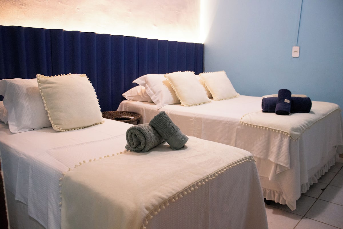 Dos camas individuales con sábanas blancas y almohadones decorativos, una mesita con una cesta y una cortina azul en una habitación de hotel.