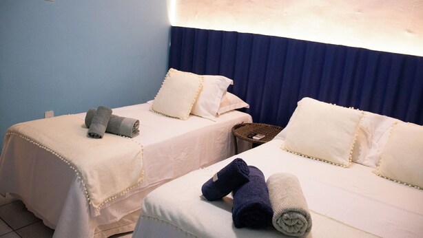 Dos camas individuales con sábanas blancas y almohadas decorativas, una mesita de mimbre pequeña y una lámpara de pared.