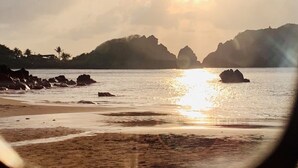 Une scène de plage avec le soleil se couchant sur l’eau, des affleurements rocheux et des collines lointaines.