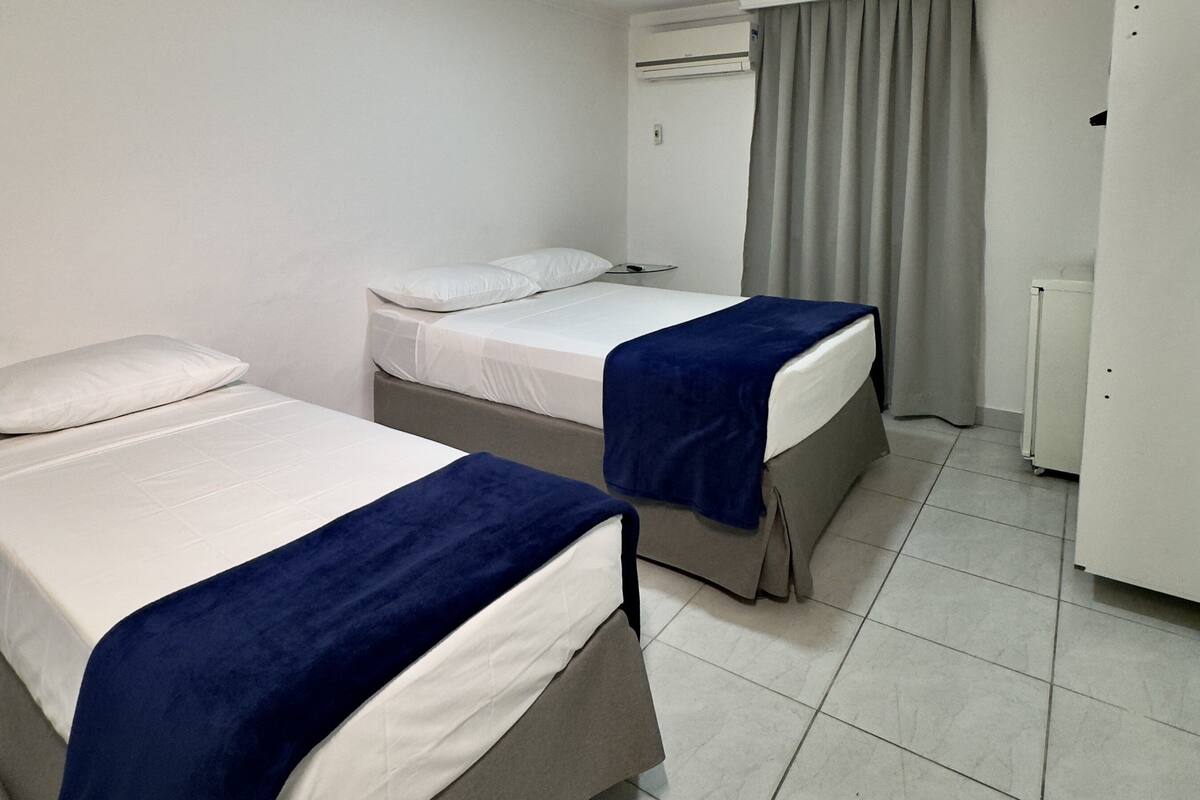 Une chambre d’hôtel avec deux lits, un luminaire au plafond, un climatiseur, une fenêtre avec des rideaux et un réfrigérateur.