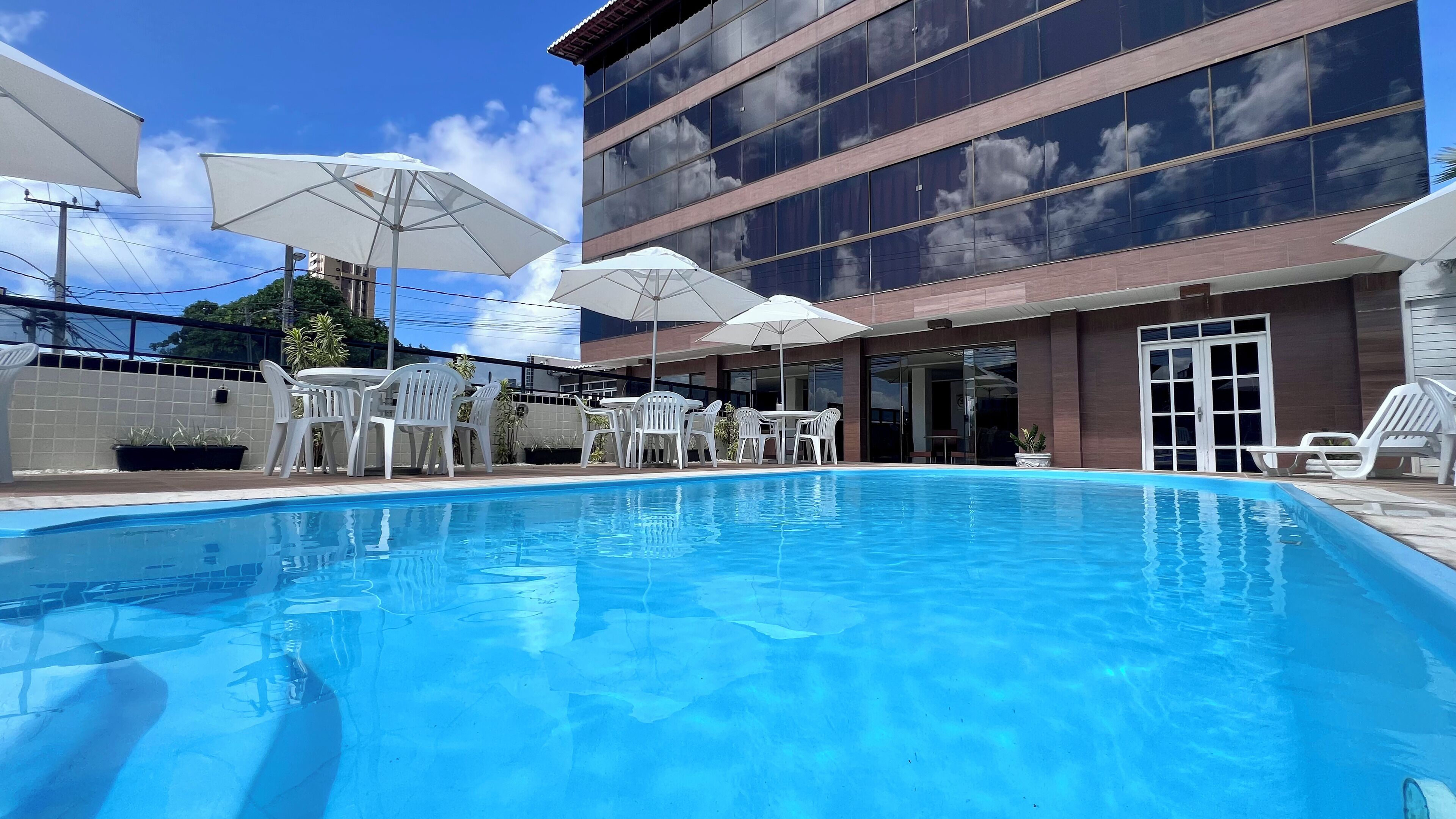 Piscina com cadeiras e guarda-sóis brancos, um edifício moderno com grandes janelas e um céu azul límpido.