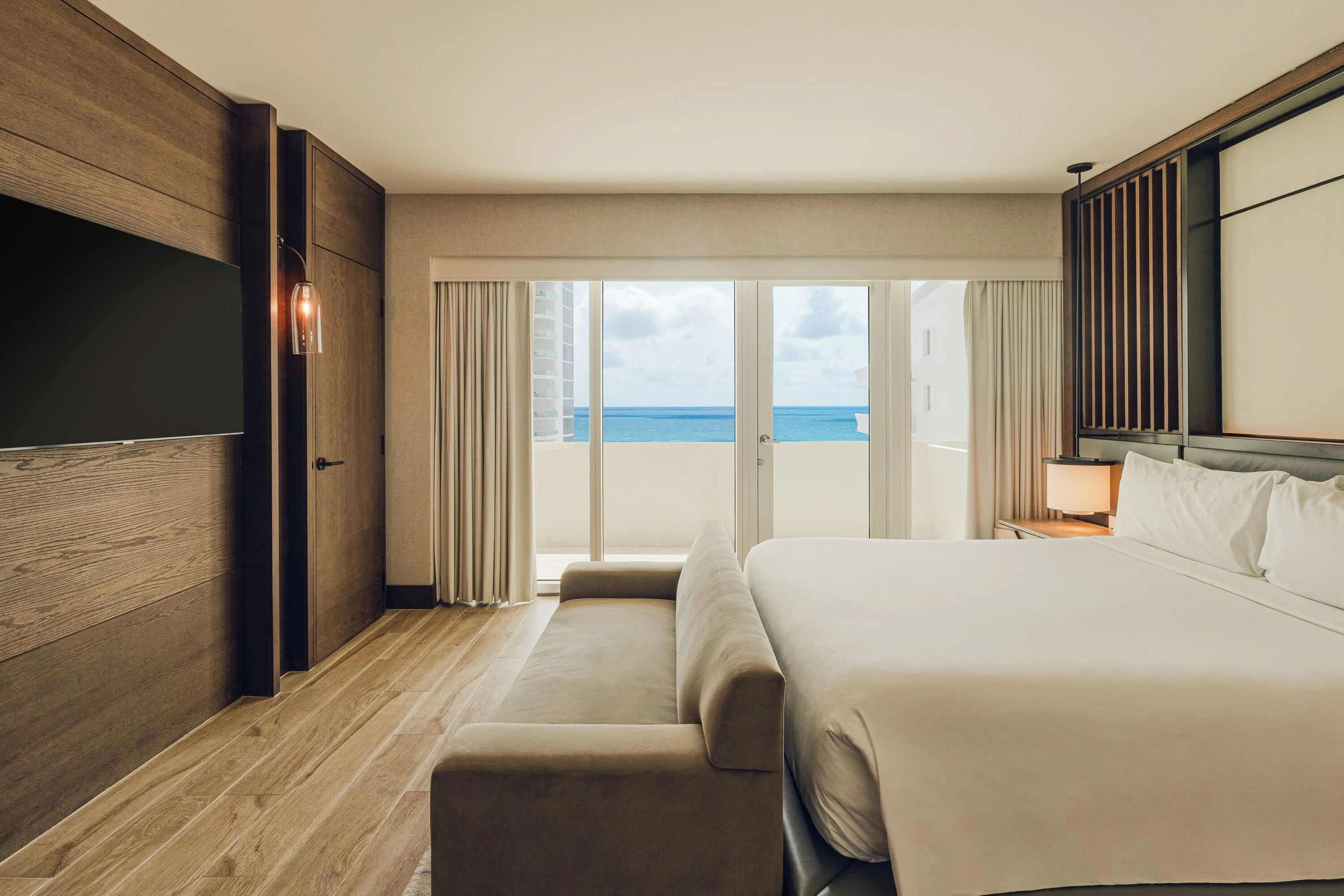 A hotel room with a bed, a TV, a balcony with a view of the sea, and a wooden floor.