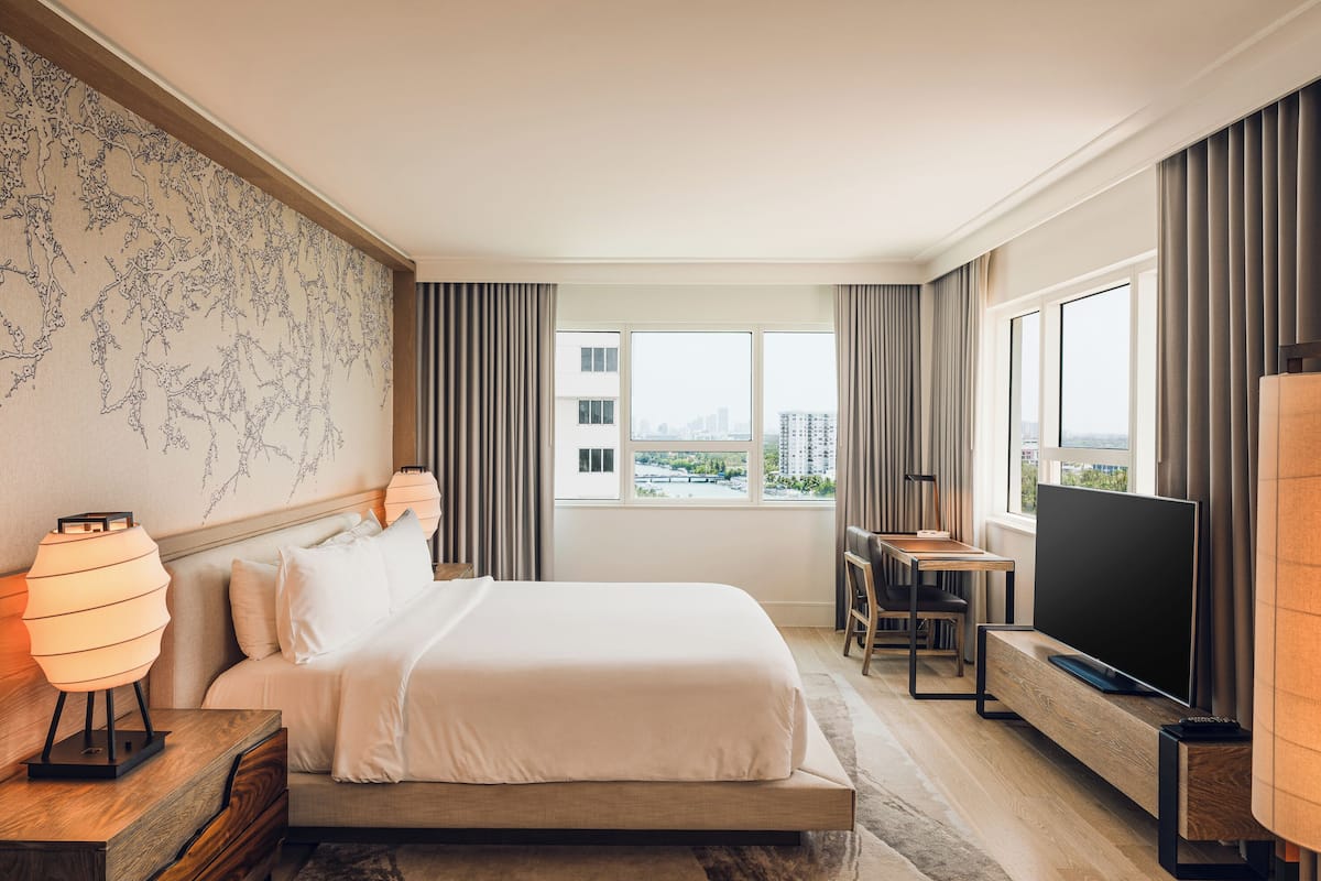 Una habitación de hotel moderna con una cama grande, un televisor de pantalla plana, un escritorio con una silla y vistas a la ciudad a través de amplias ventanas.