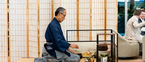 Una persona seduta su un tatami di fronte a un tradizionale paravento giapponese, con un tavolino su cui è posata una teiera e accessori per il tè.