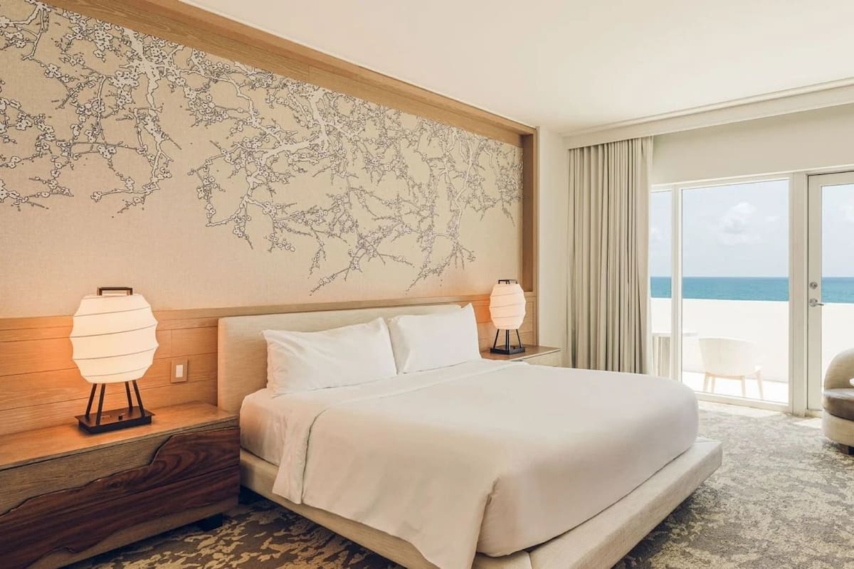 Habitación de hotel con cama, sofá, una mesita, un ventanal con vista al mar y una pared decorada con un motivo de árbol.