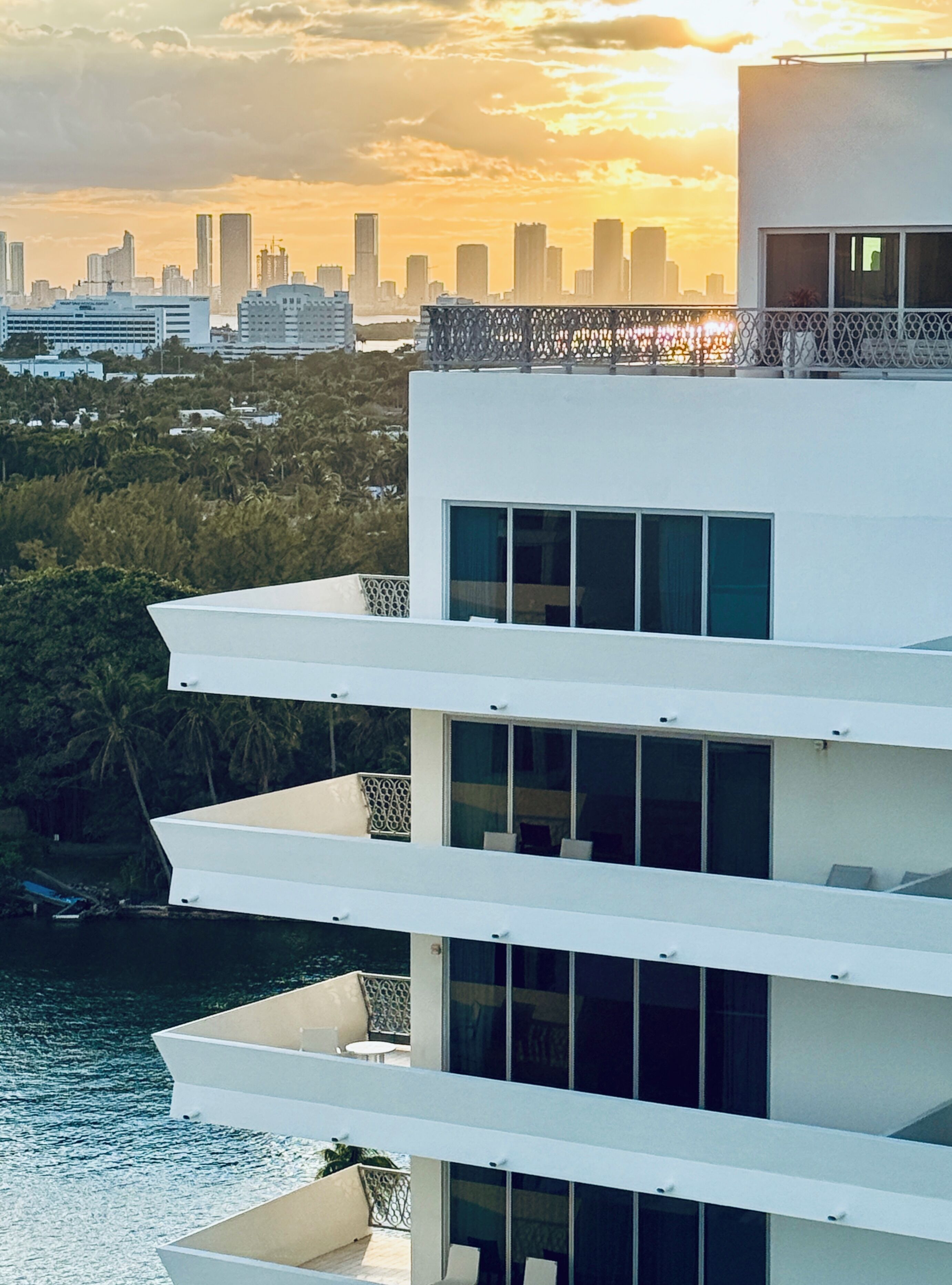 Un edificio moderno de varios pisos con balcones que dan a una masa de agua y un horizonte urbano al fondo.