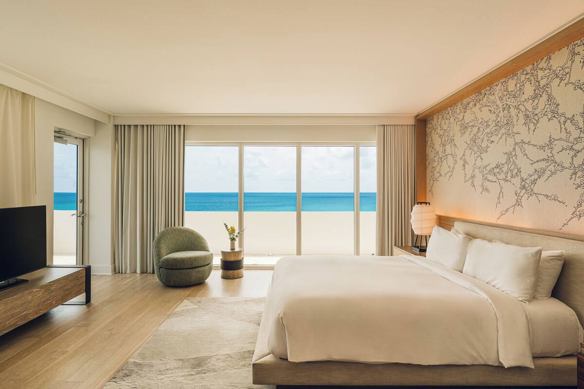 Una habitación de hotel moderna con una cama grande, televisión, un área de descanso con una silla y vista al océano.