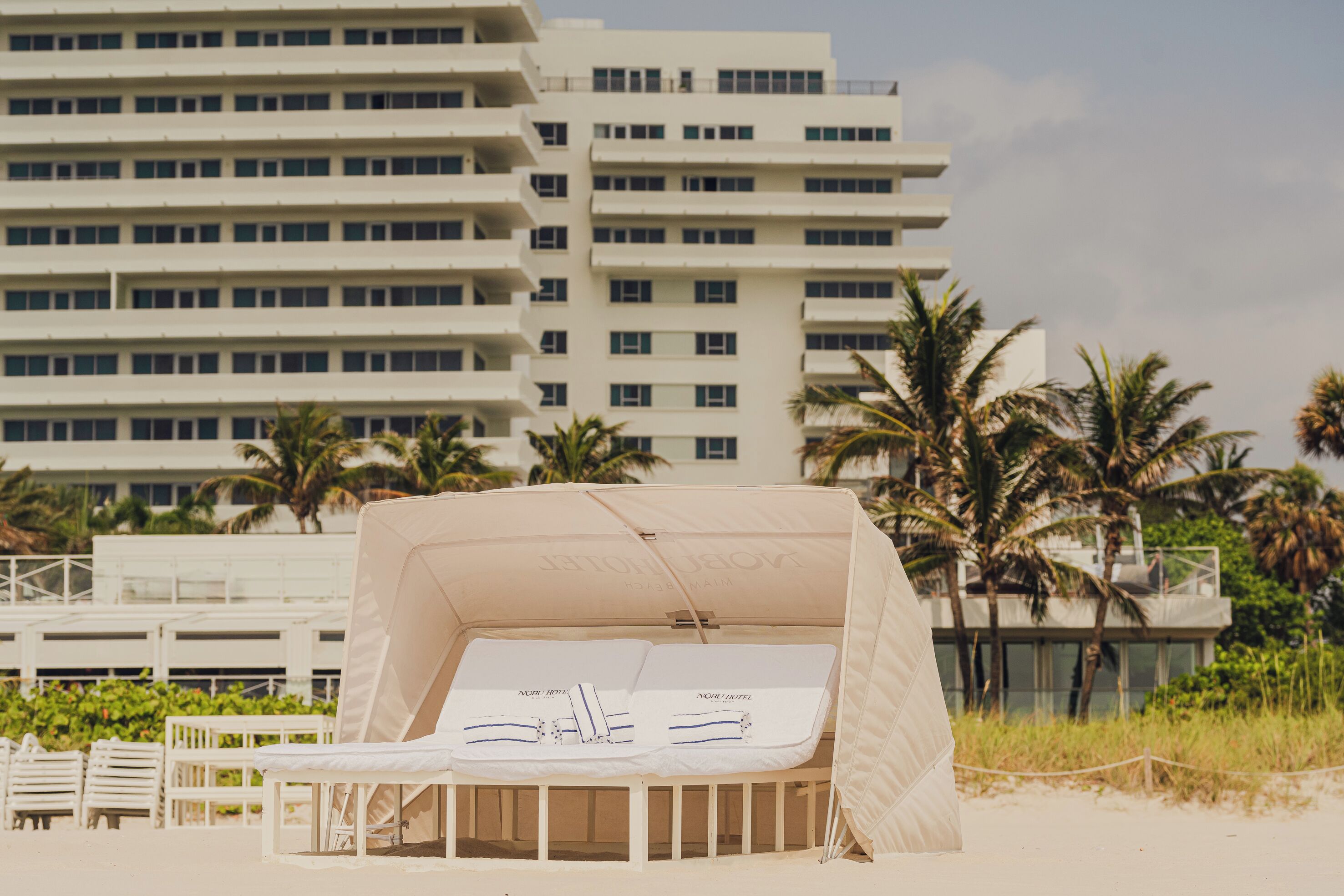 A beach cabana with a canopy, white cushions, and a small table. There are palm trees and a multi-story building in the background.