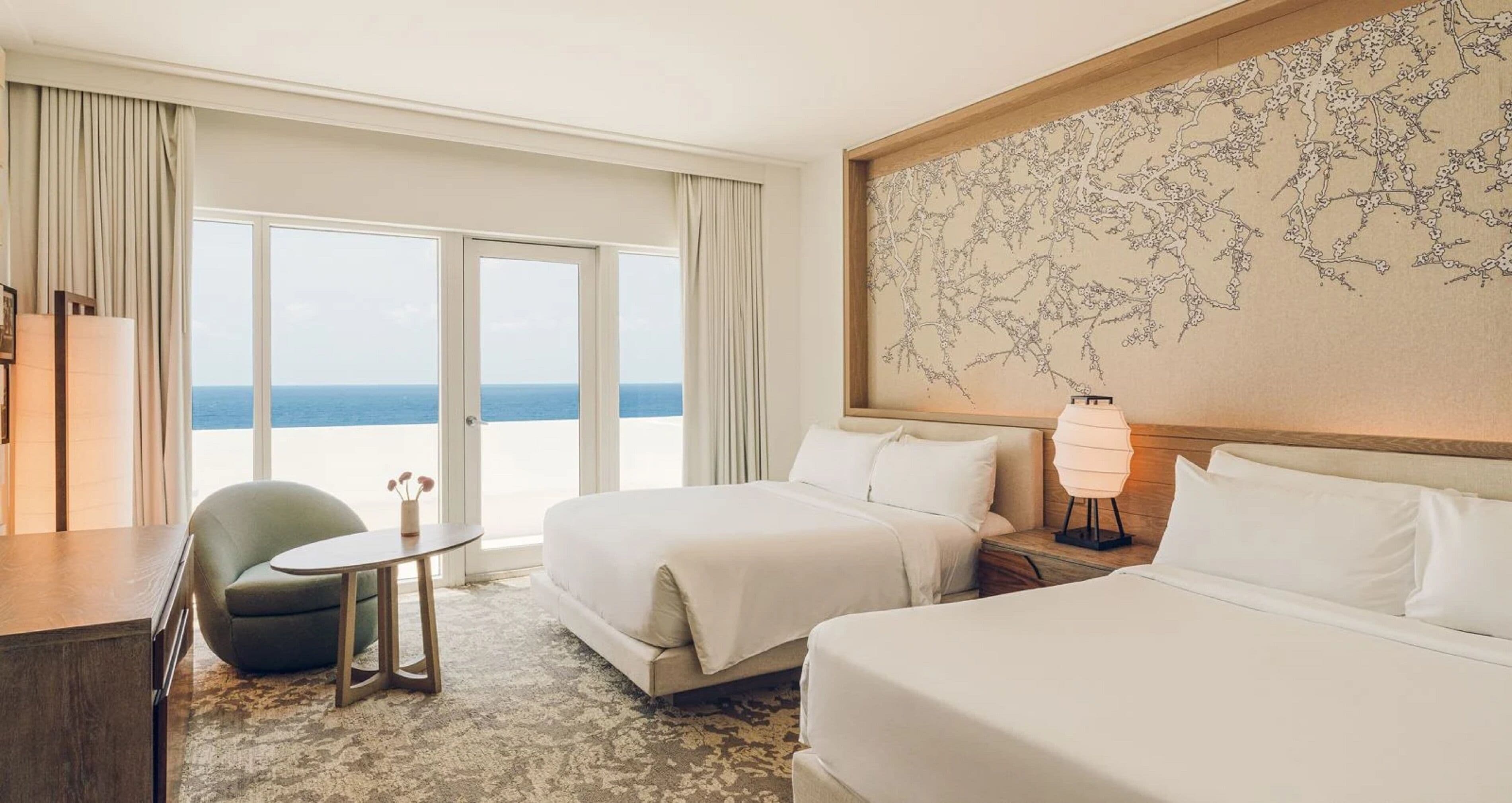 A hotel room with two beds, a desk, a chair, a lamp, and a large window with a view of the sea.