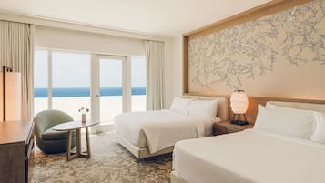 Habitación de hotel con dos camas, un escritorio, una silla, una lámpara y un ventanal con vistas al mar.
