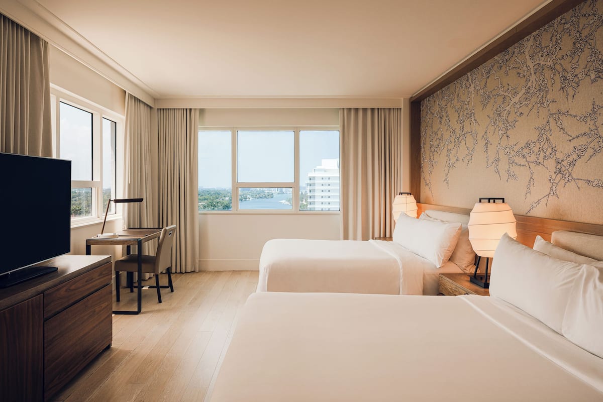 Habitación de hotel con dos camas, un escritorio con silla, televisión y un ventanal con vistas a la ciudad.