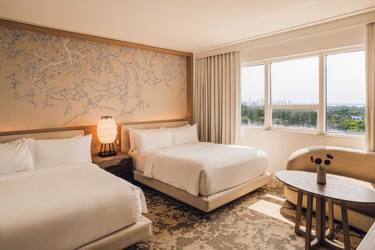 Habitación de hotel con dos camas, una mesita de noche con lámpara, una pequeña mesa redonda con un jarrón y vistas a la ciudad a través de amplias ventanas.