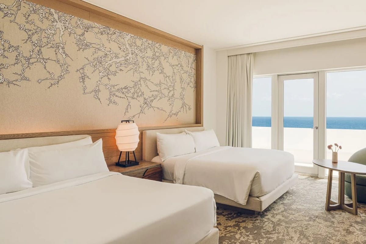 Habitación de hotel con dos camas, un escritorio, una silla, una lámpara y vista al océano.