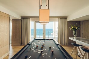 A pool table in a room with a view of the sea, wooden cabinets, and a bar area.