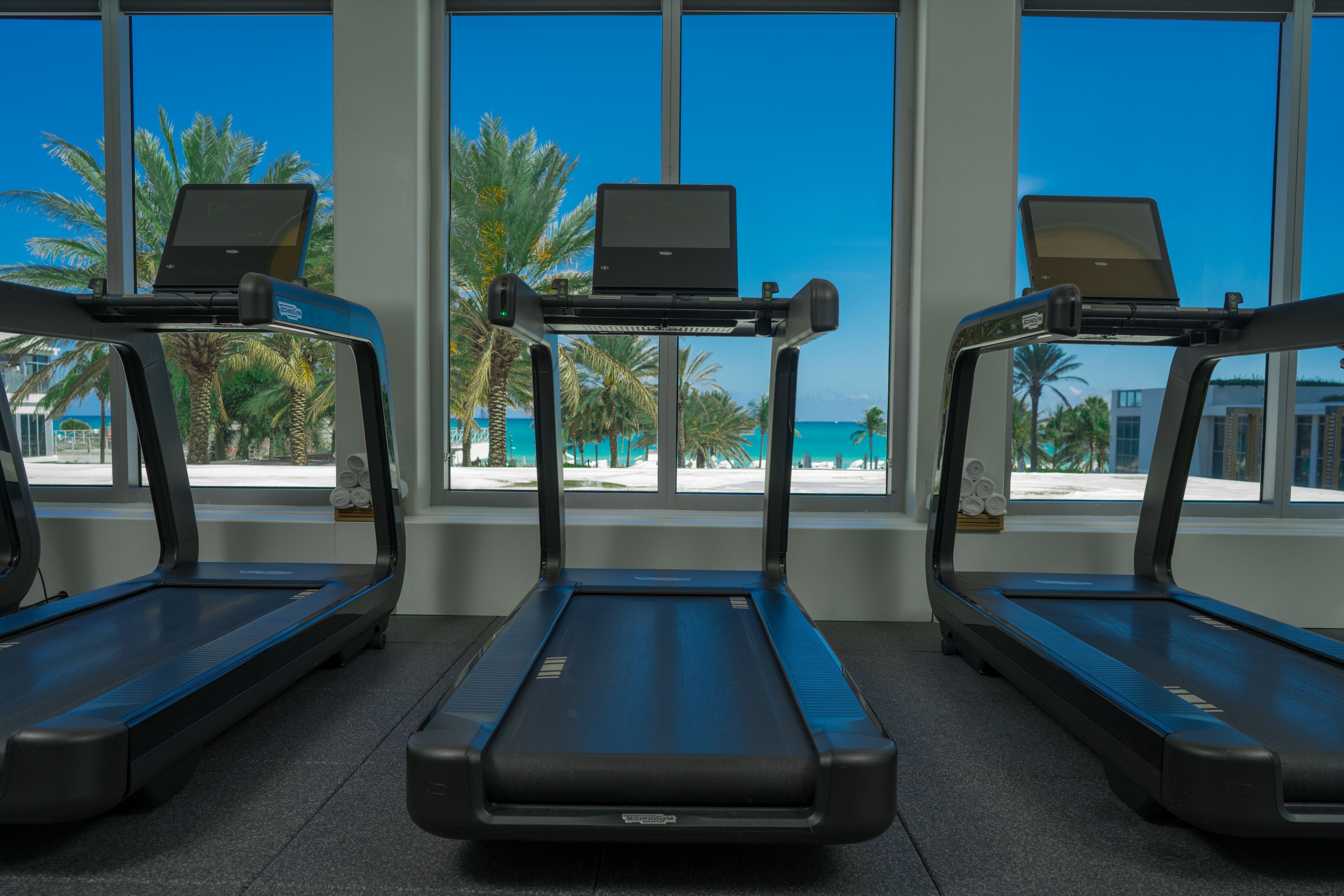 Three treadmills with laptops on them, positioned in front of large windows with a view of palm trees and a beach.