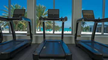 Three treadmills with laptops on them, positioned in front of large windows with a view of palm trees and a beach.