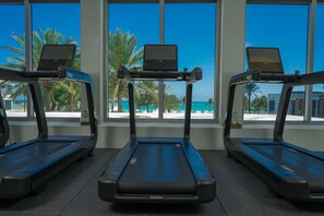 Three treadmills with laptops on them, positioned in front of large windows with a view of palm trees and a beach.