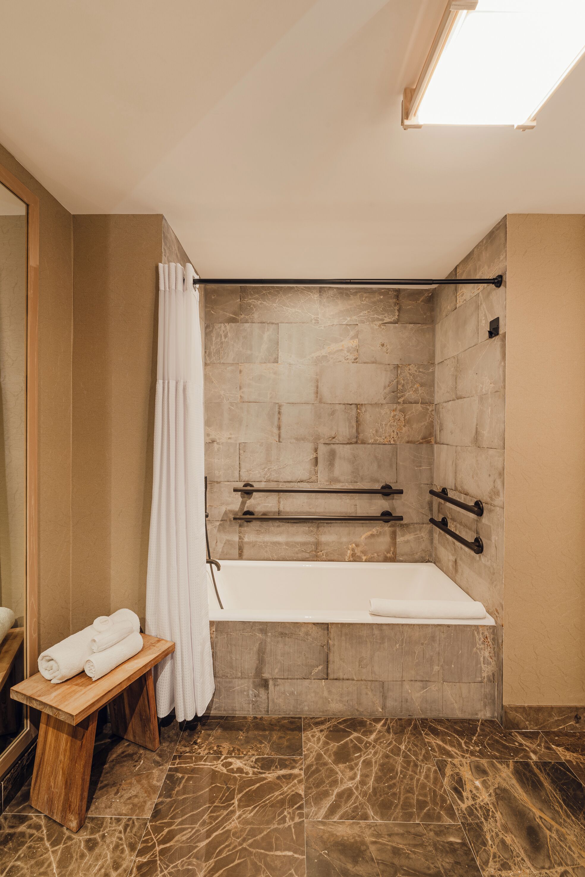 A modern bathroom with a stone wall, a bathtub, a wooden bench with towels, and a glass shower enclosure.