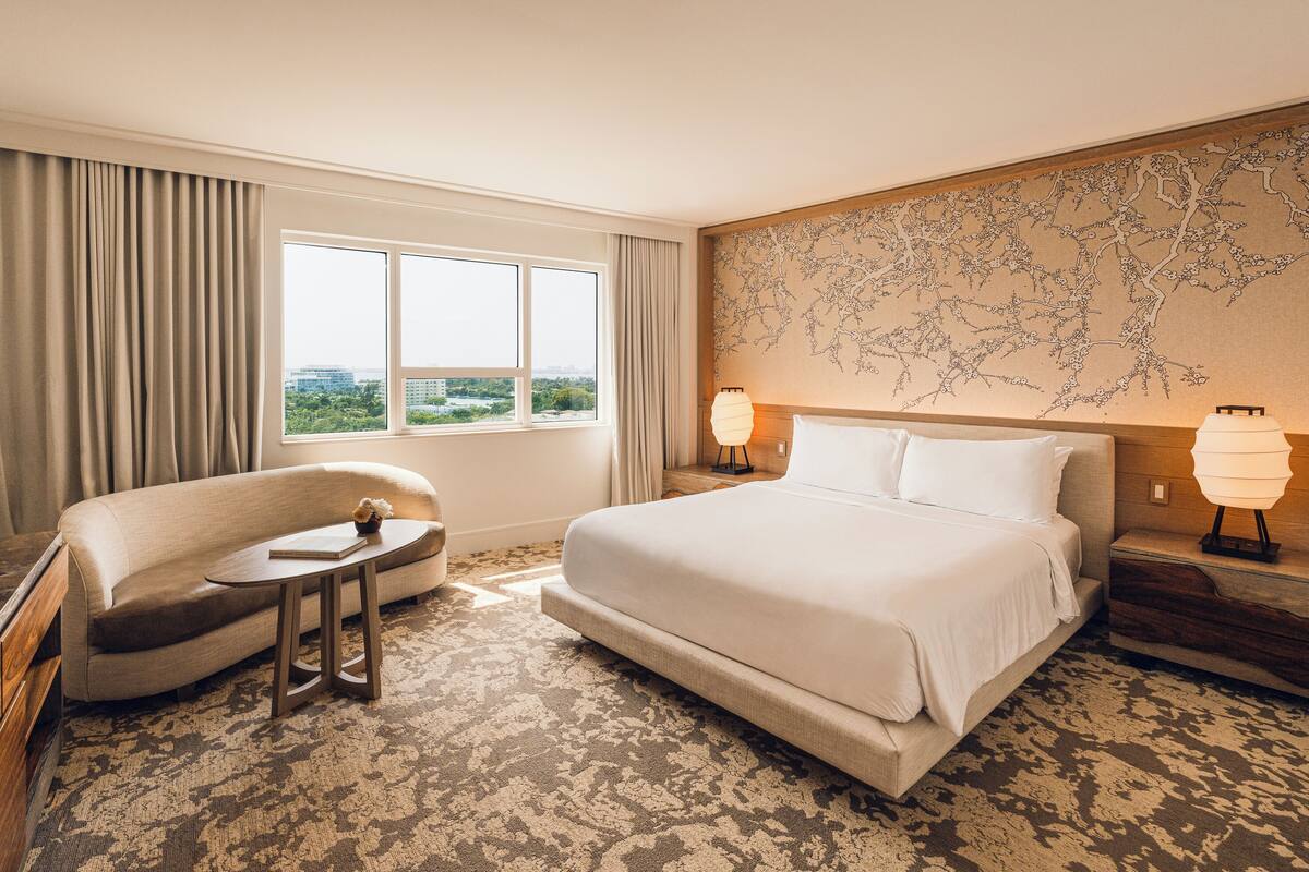 Habitación de hotel con una cama grande, un sofá, una mesita y vistas a la ciudad por la ventana.