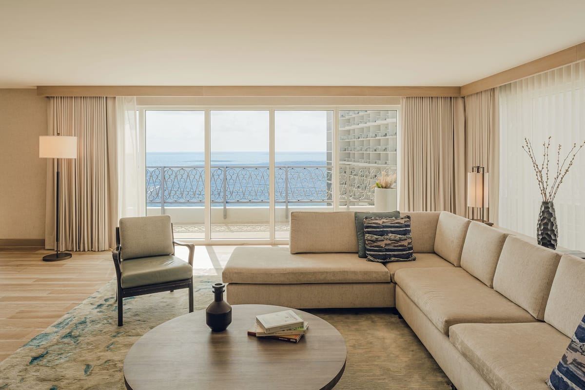 Habitación de hotel con un ventanal grande, un sofá, una silla, una mesita de centro y una lámpara.