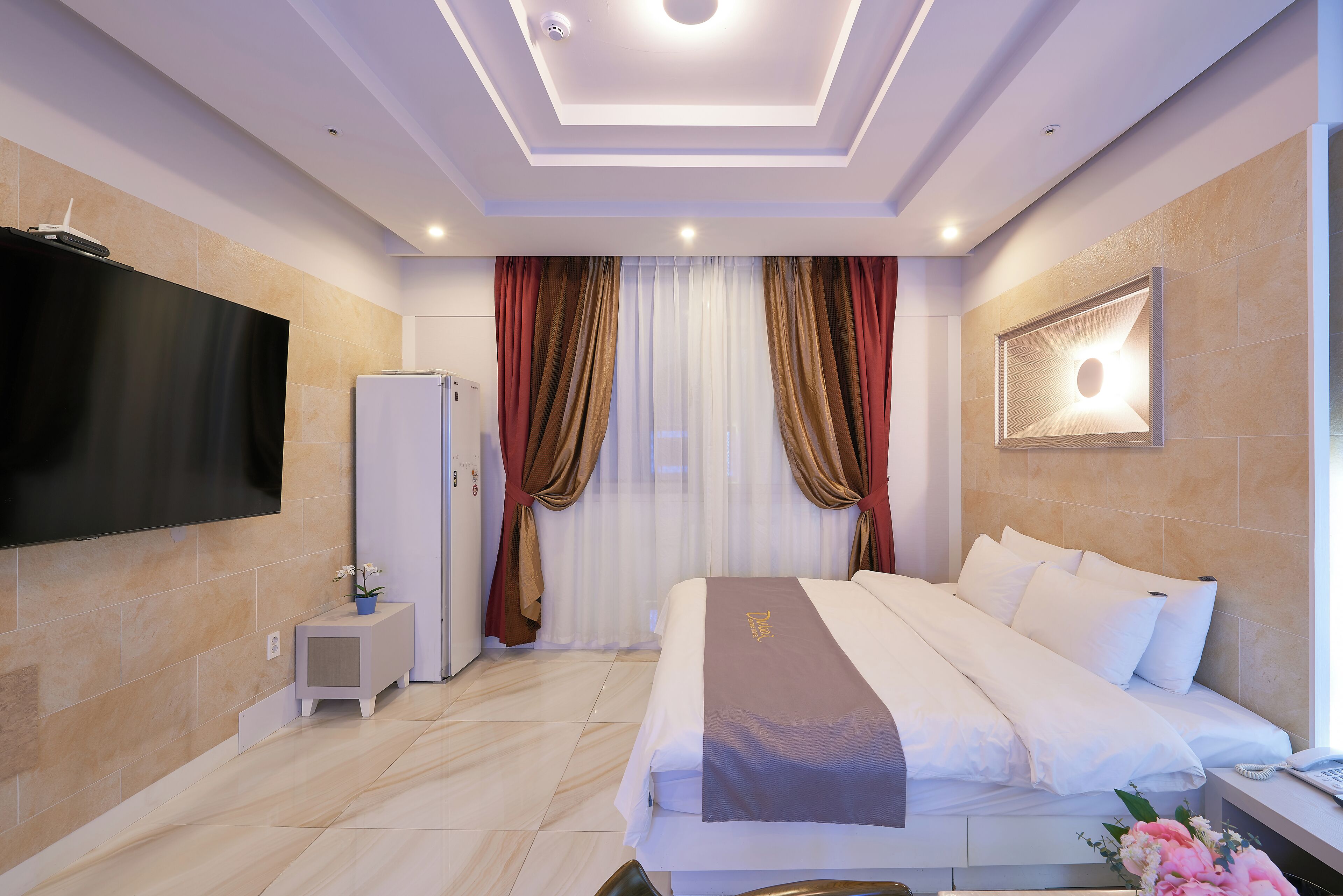 A modern hotel room with a large bed, a flat-screen TV, a minibar, and a large window with curtains.