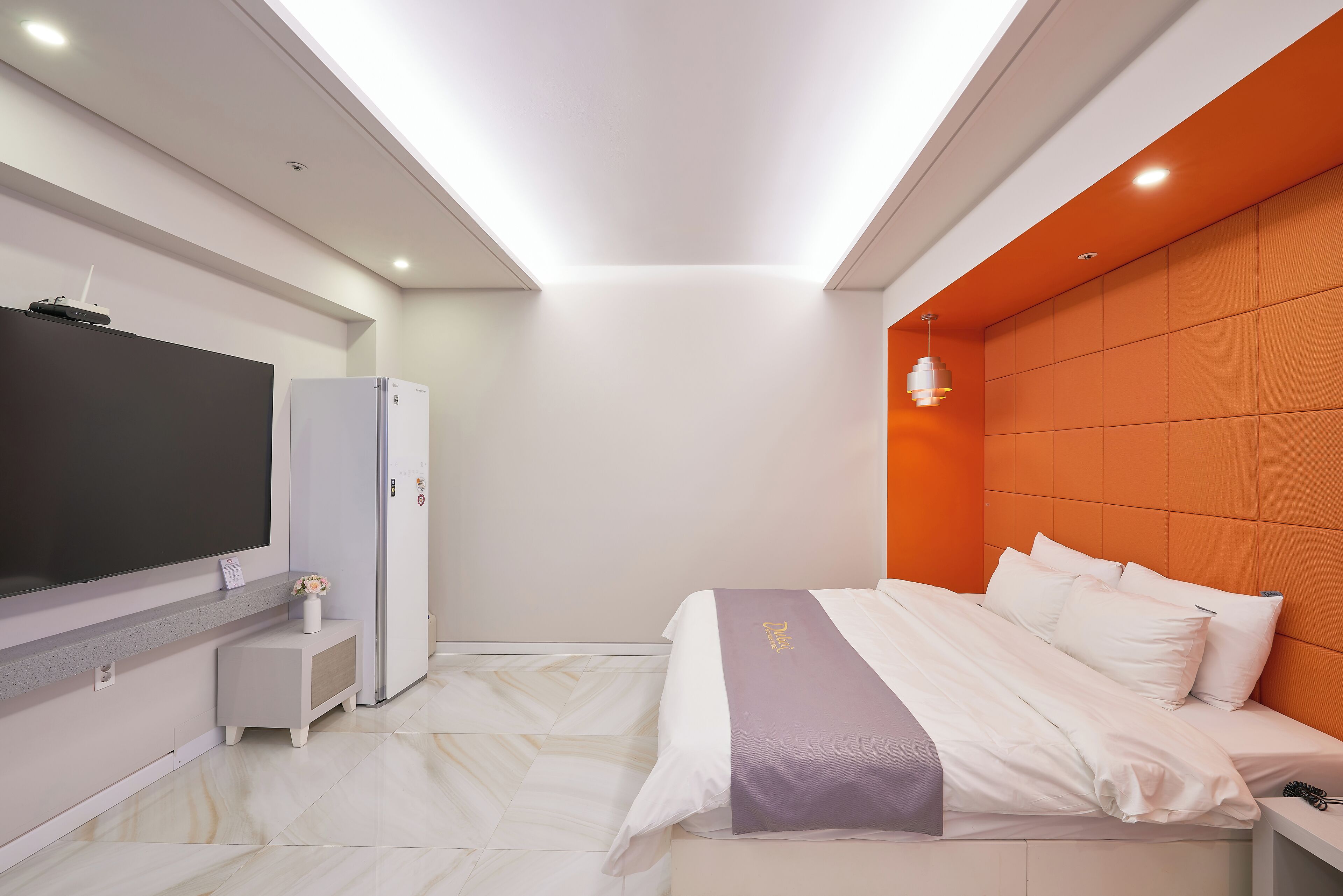 A modern hotel room with a large flat-screen TV, a mini-fridge, a bedside table with a vase, and a bed with a purple throw.