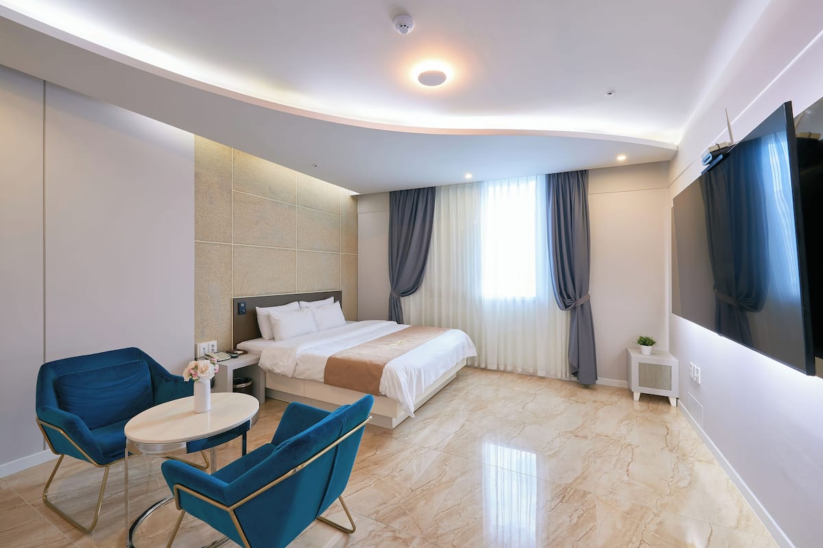 Quarto de hotel moderno com uma cama, duas poltronas azuis, uma mesa pequena e uma televisão.
