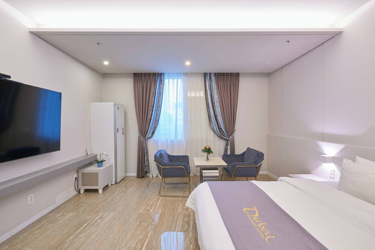 Quarto de hotel com cama, televisão, escrivaninha com duas cadeiras, frigorífico e cortinas.