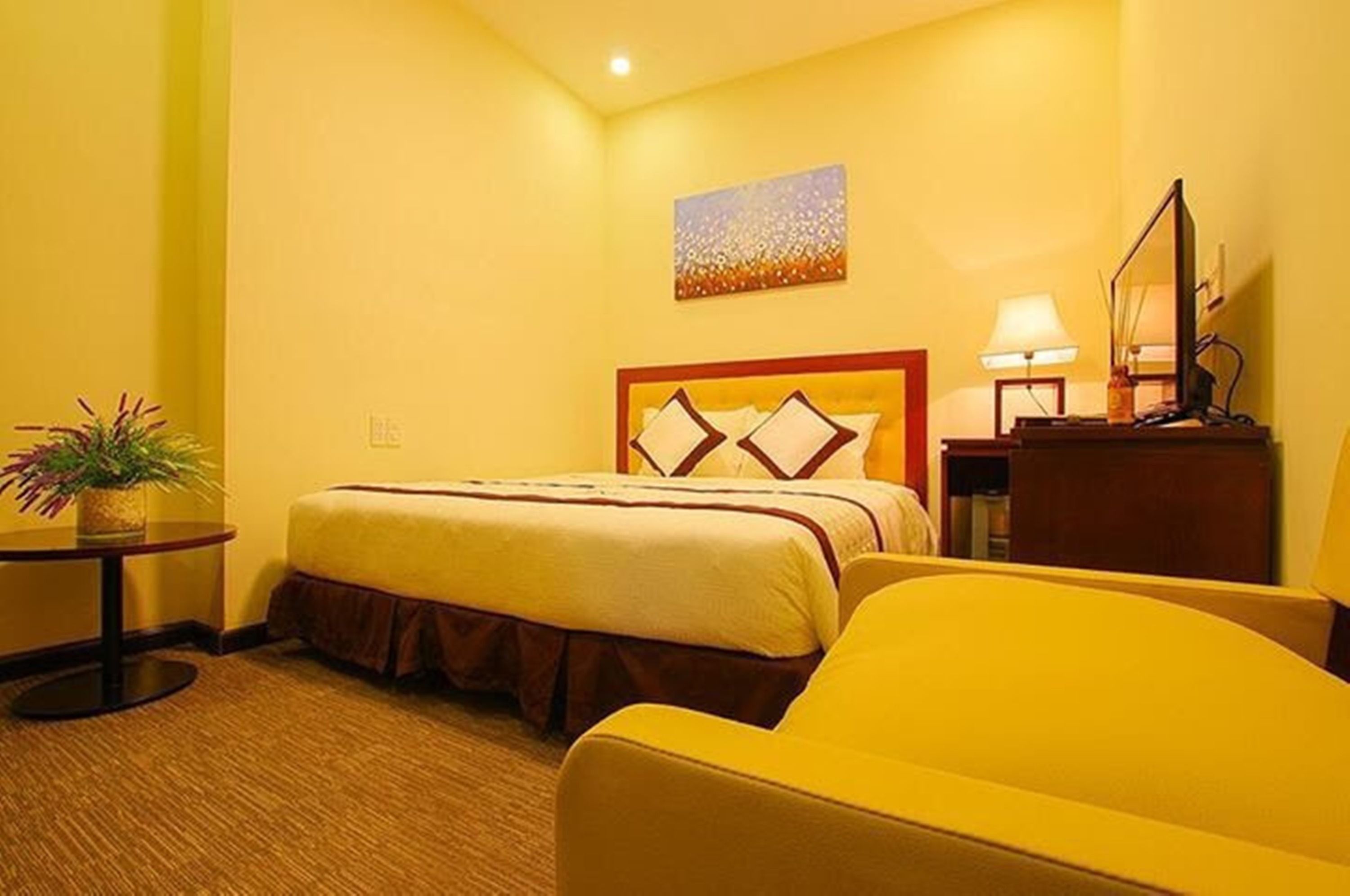 A hotel room with a bed, a chair, a small table with a plant, a lamp, and a painting on the wall.