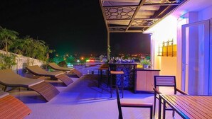 Una terraza en la azotea con sillones, una zona de bar y vistas a la ciudad de noche.