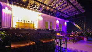 Una zona de bar con taburetes y mesas de madera, una barra con botellas y un techo decorativo con un diseño geométrico.