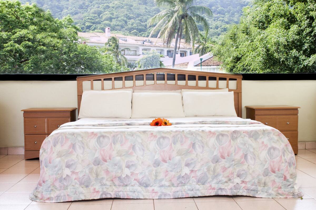 A bedroom with a bed, two nightstands, and a view of a lush green landscape with trees and a building in the background.