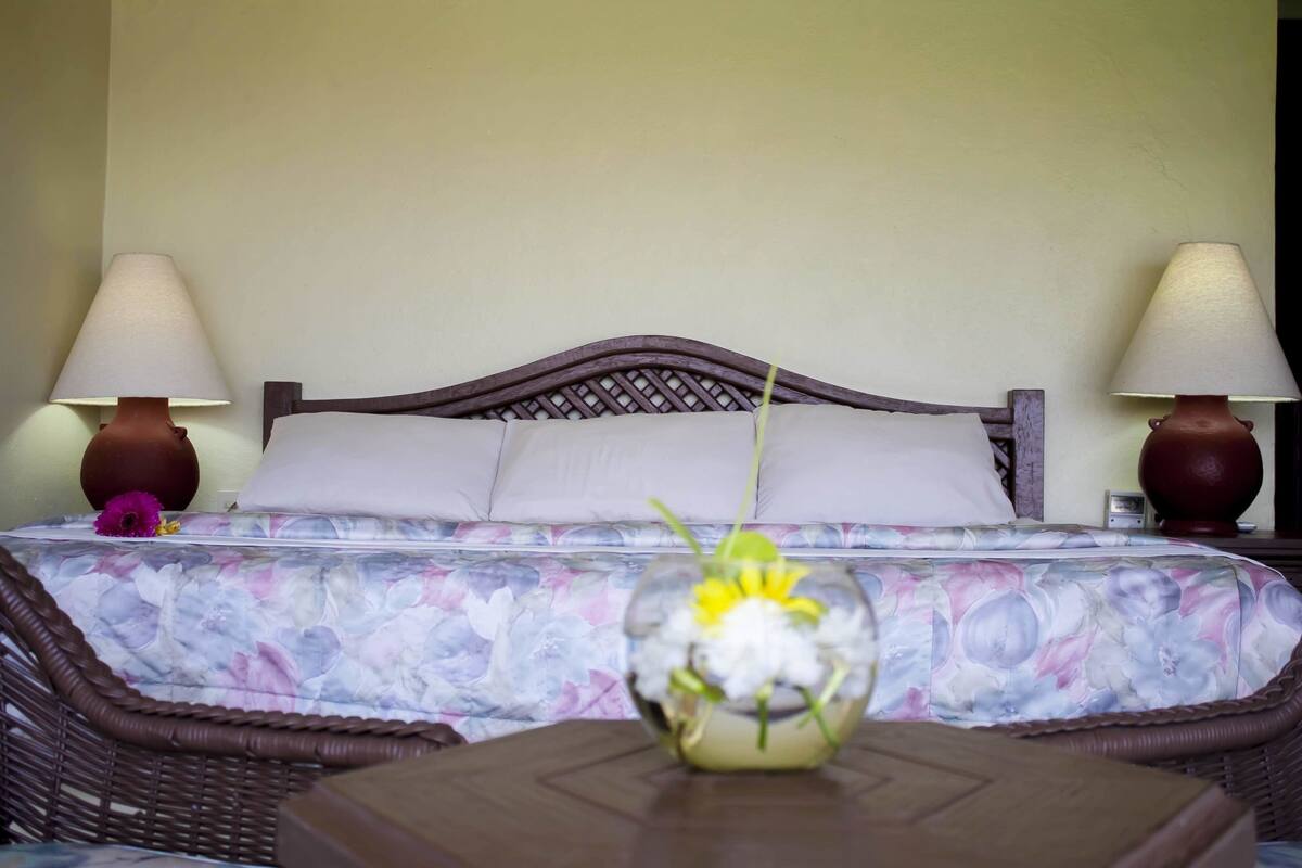 A hotel room with a bed, two bedside lamps, a floral bedspread, and a vase with flowers on a table.