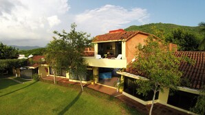 A two-story house with a balcony, surrounded by greenery and hills in the background.