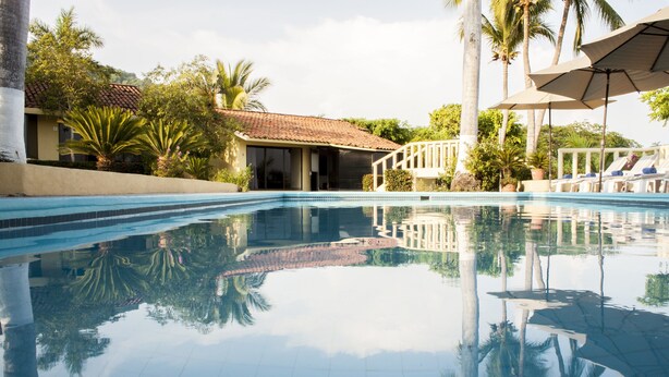 A swimming pool with tiled edges, surrounded by palm trees and a building with a tiled roof.