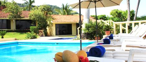Zona de piscina con sillas de descanso, una mesa con frutas y una flor, y un edificio con techo de tejas.