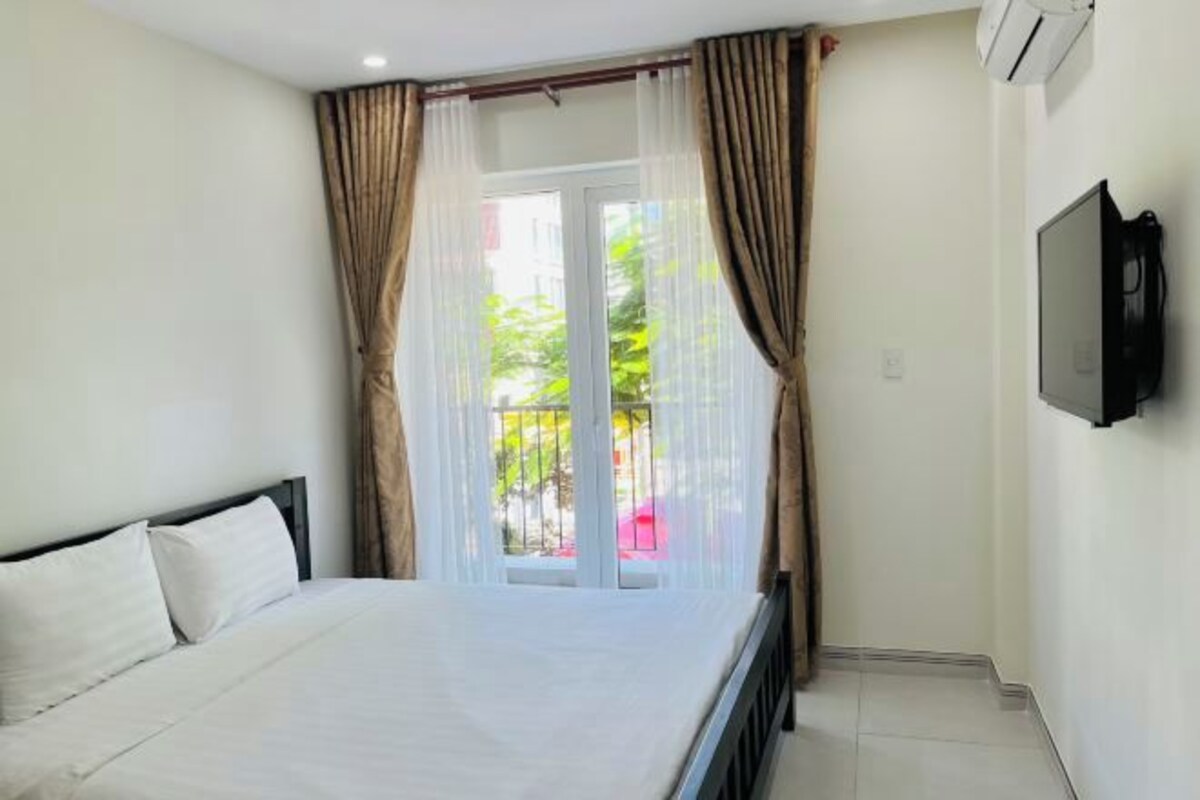 A hotel room with a bed, curtains, a TV, and a view of greenery outside.