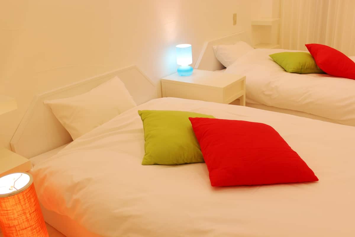 Habitación de hotel con dos camas, cada una con ropa de cama blanca y almohadas de colores. Se observan mesitas de noche con lámparas y una pequeña mesa al lado de cada cama.