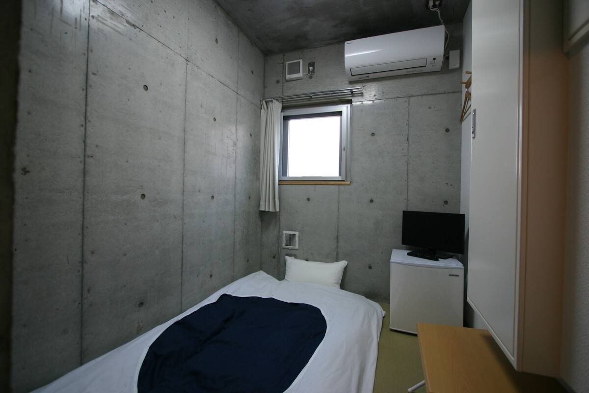 A small, compact room with a bed, a desk, a television, and an air conditioning unit.